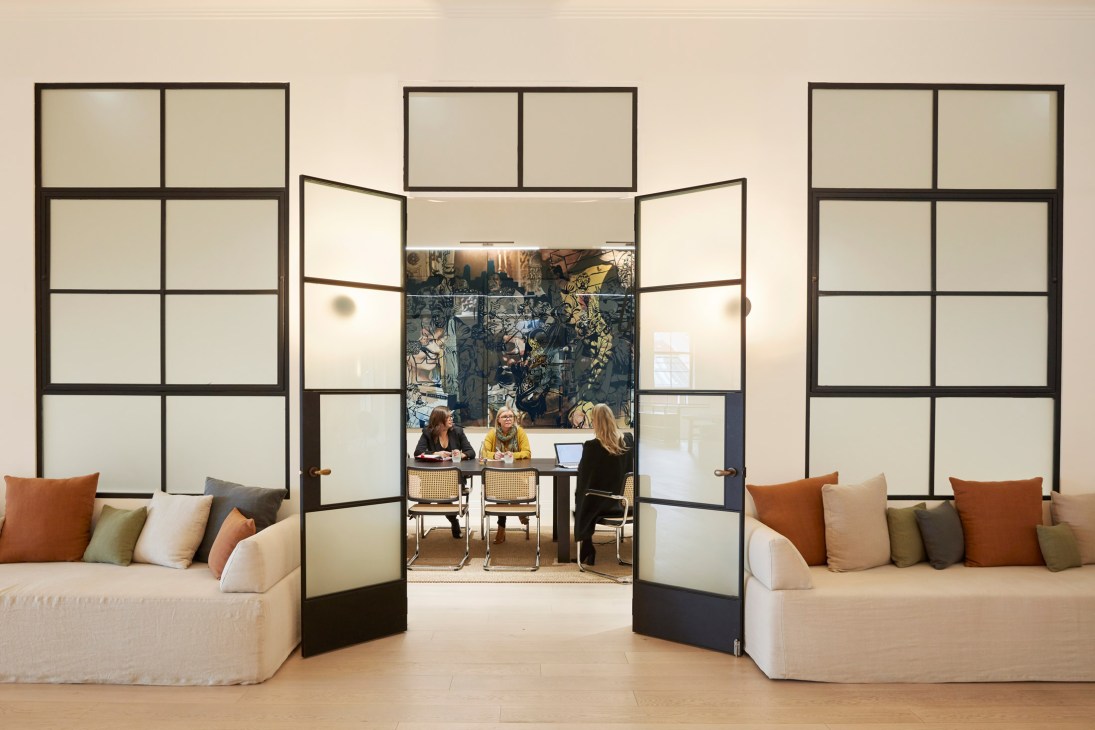 Modern office space with glass partition doors opening to a meeting room where three people sit at a table, flanked by cream sofas with cushions.