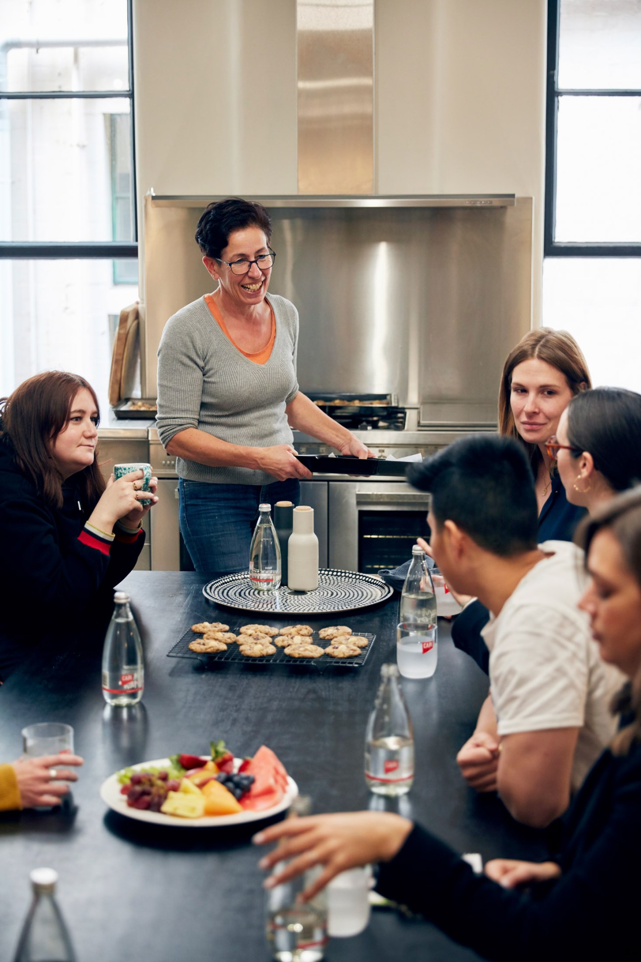 A woman stands at a kitchen counter serving food to seated colleagues during a shared lunch at Capi's office headquarters.