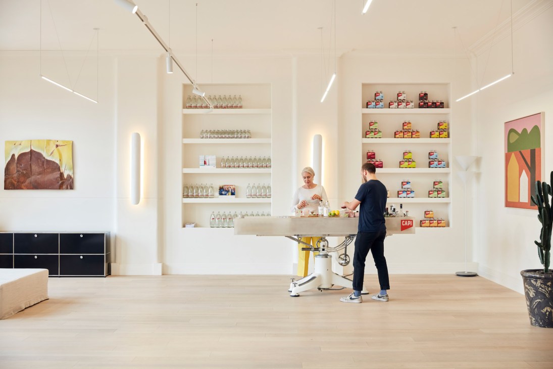 Two people preparing cocktails at a minimalist bar counter with shelves of Capi bottles displayed behind them.