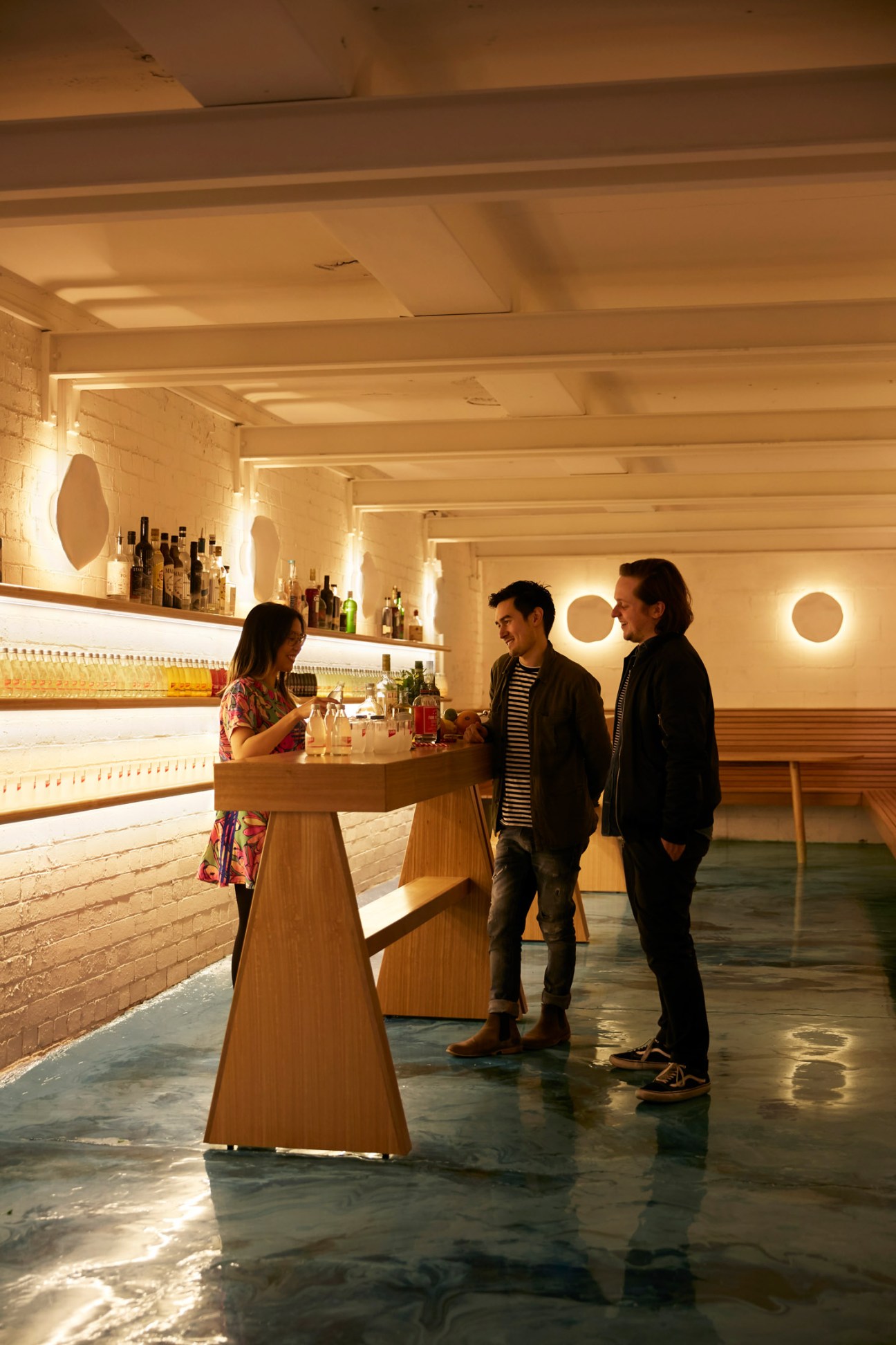 A woman bartender serves drinks to two men at a modern wooden bar with illuminated circular wall features and bottle displays.