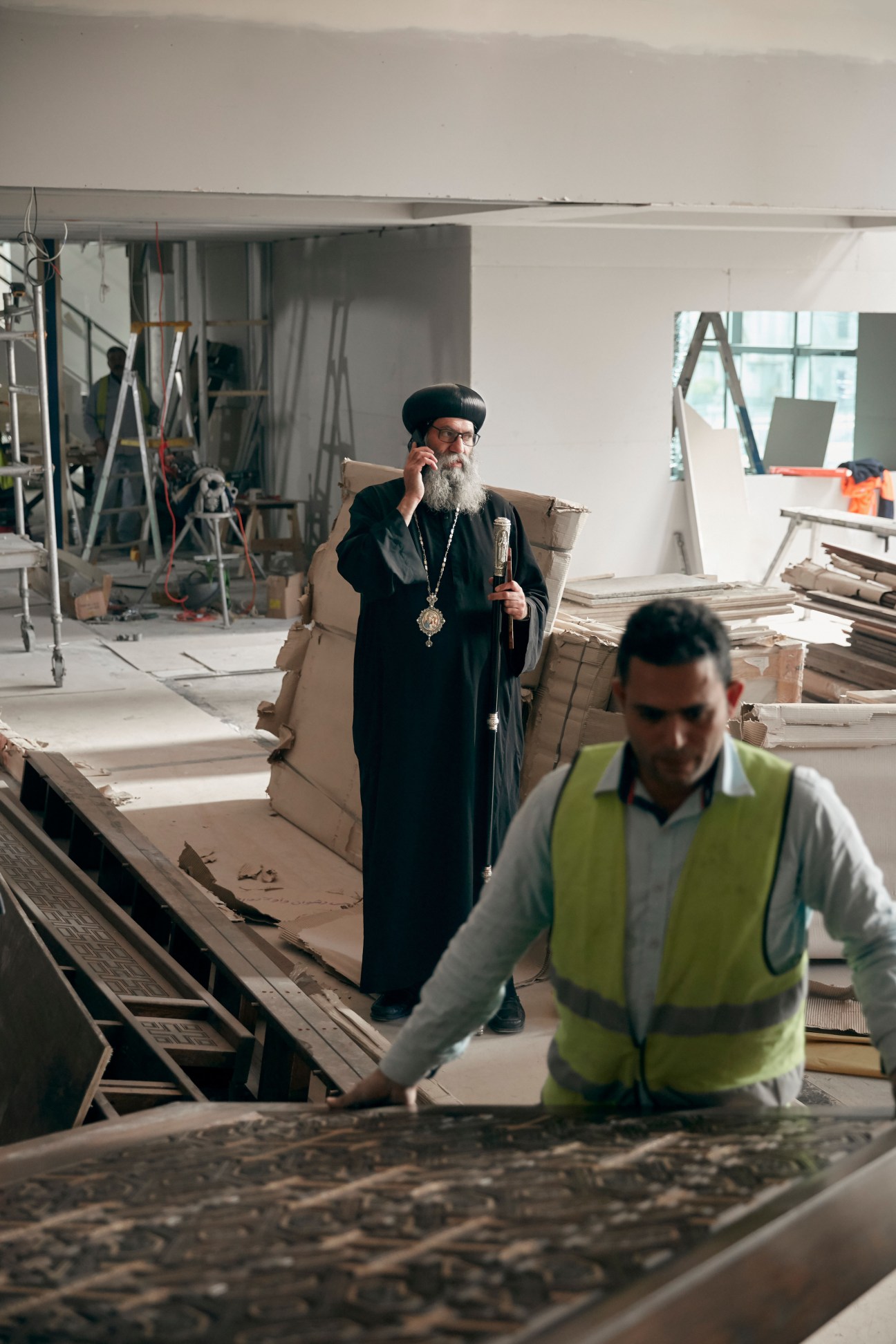 Bishop Suriel in black robes and cross necklace supervises construction work on an ornate panel at Eporo Tower.