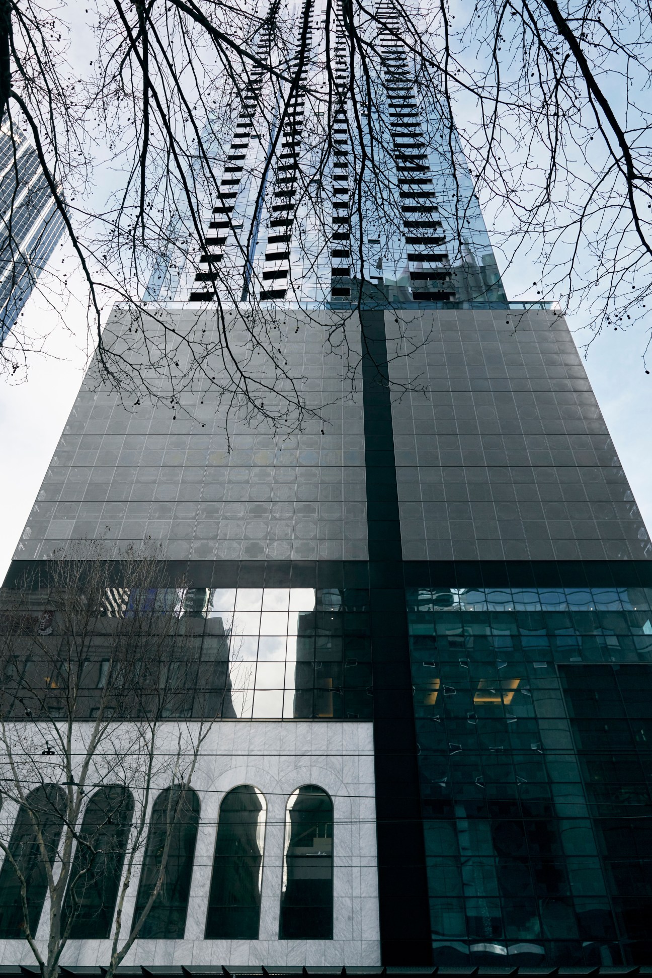 Melbourne's Eporo Tower viewed from street level, showing modern glass and perforated metal facade with bare tree branches in foreground.