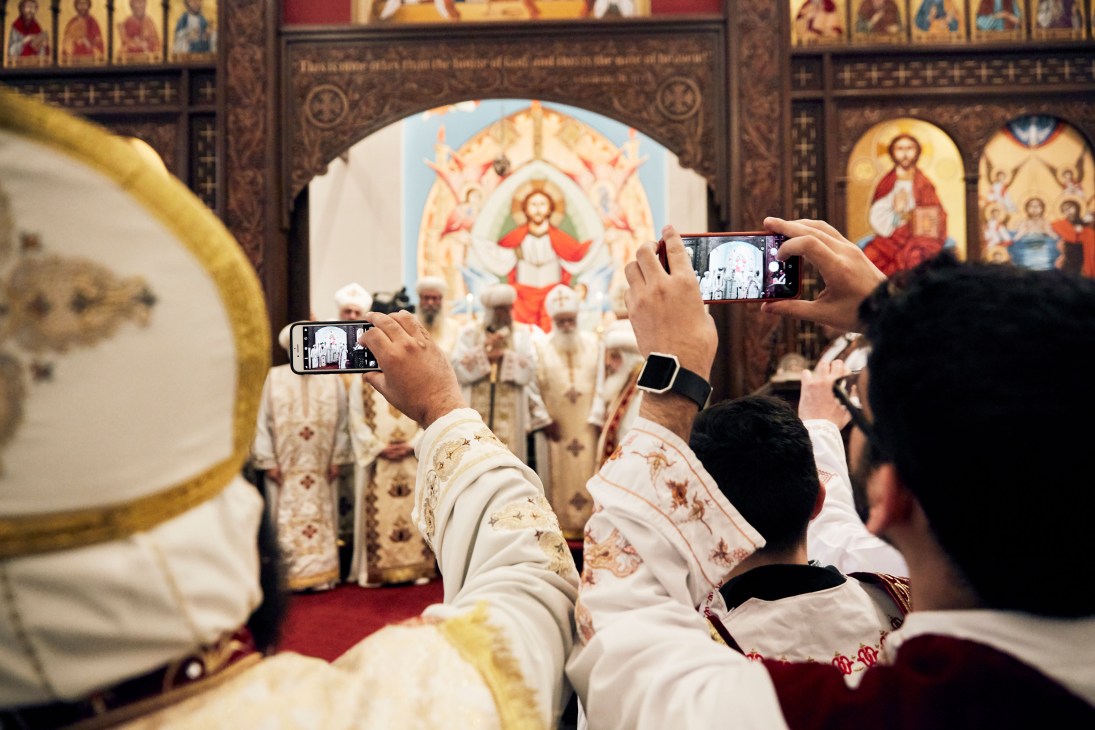 Congregation members holding up smartphones to photograph clergy during a Coptic Orthodox religious ceremony.