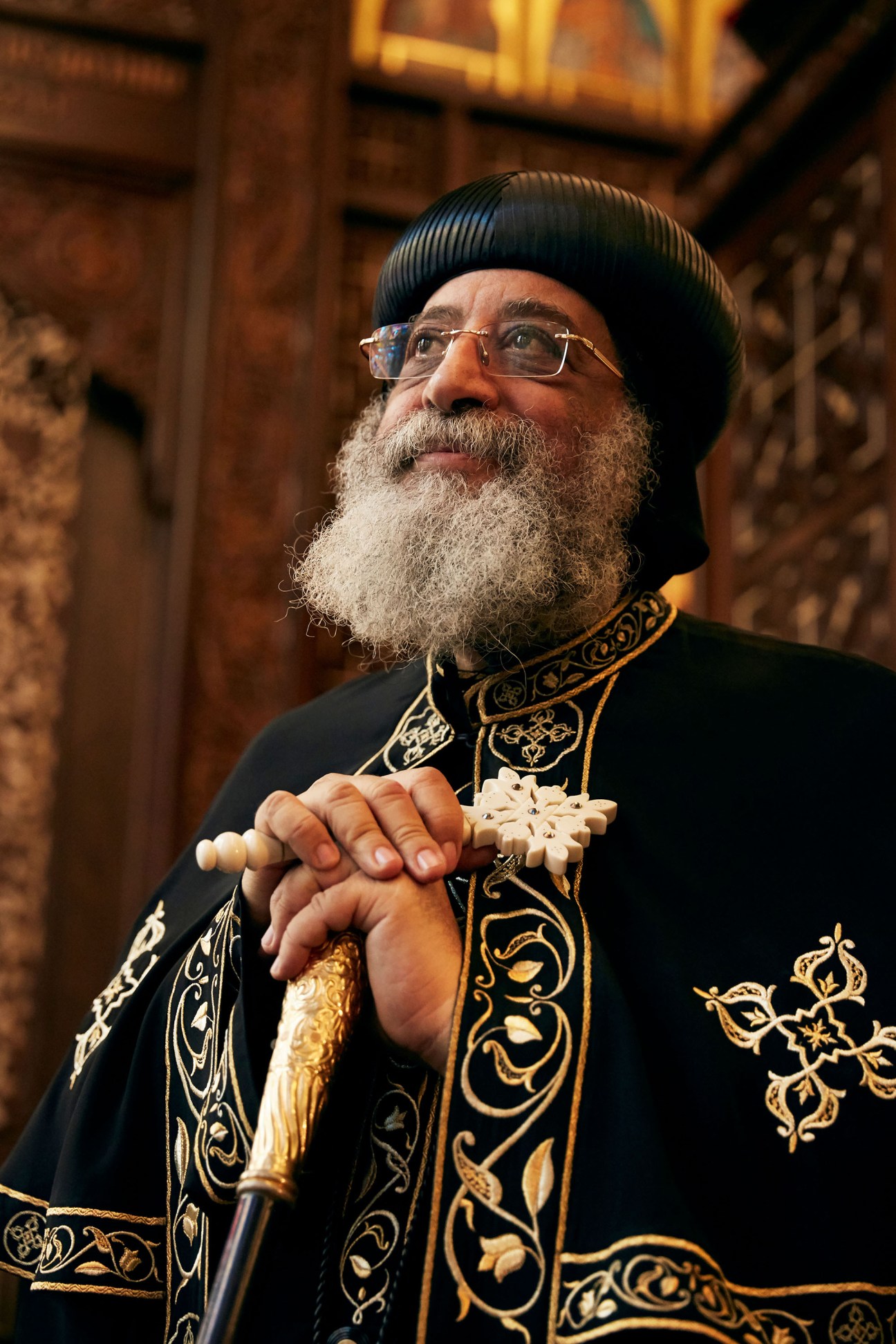 Pope Tawadros II wearing traditional Coptic Orthodox ceremonial robes with ornate gold embroidery and a black cap, holding a ceremonial staff.