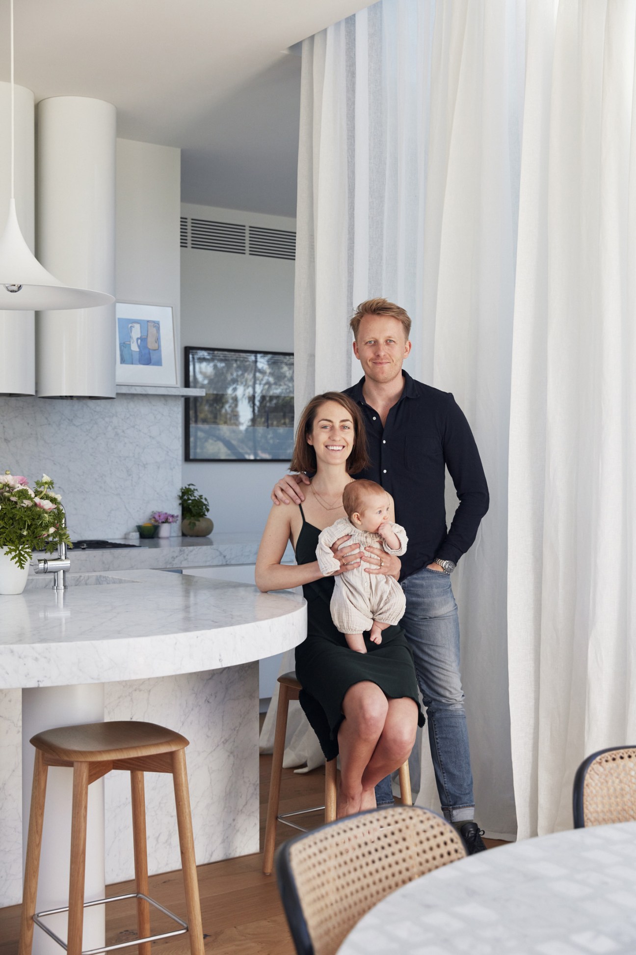 Olivia Donati-Beech stands with her husband and baby daughter in a minimalist kitchen with marble countertops and large windows.