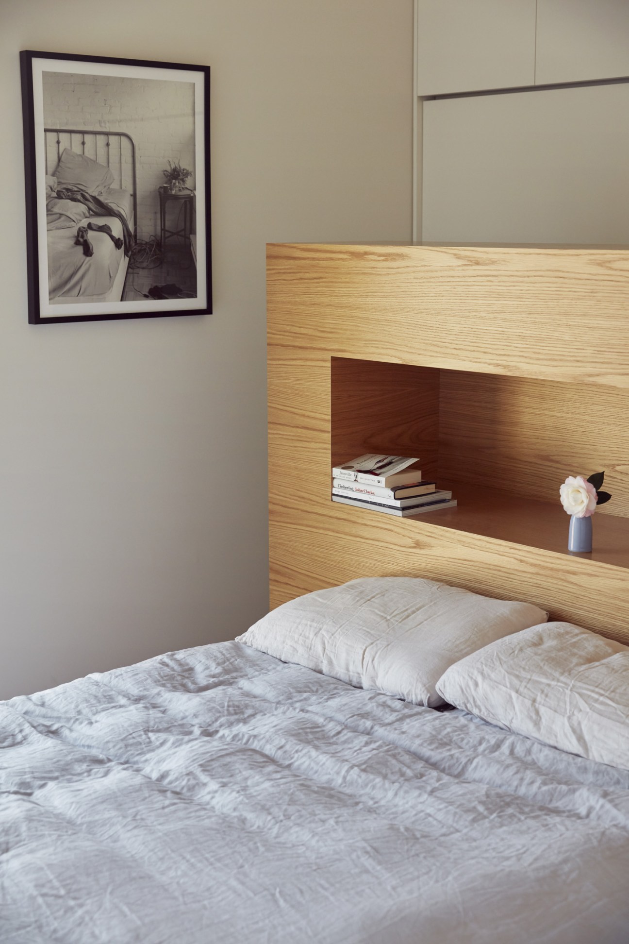 A minimalist bedroom with wooden built-in headboard featuring open shelving, white bedding, and framed artwork on pale walls.