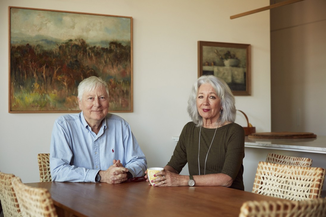 Malcolm Good and Betsy Bradshaw sit at a wooden table in their Whitlam Place apartment, with framed landscape artwork on the wall behind them.