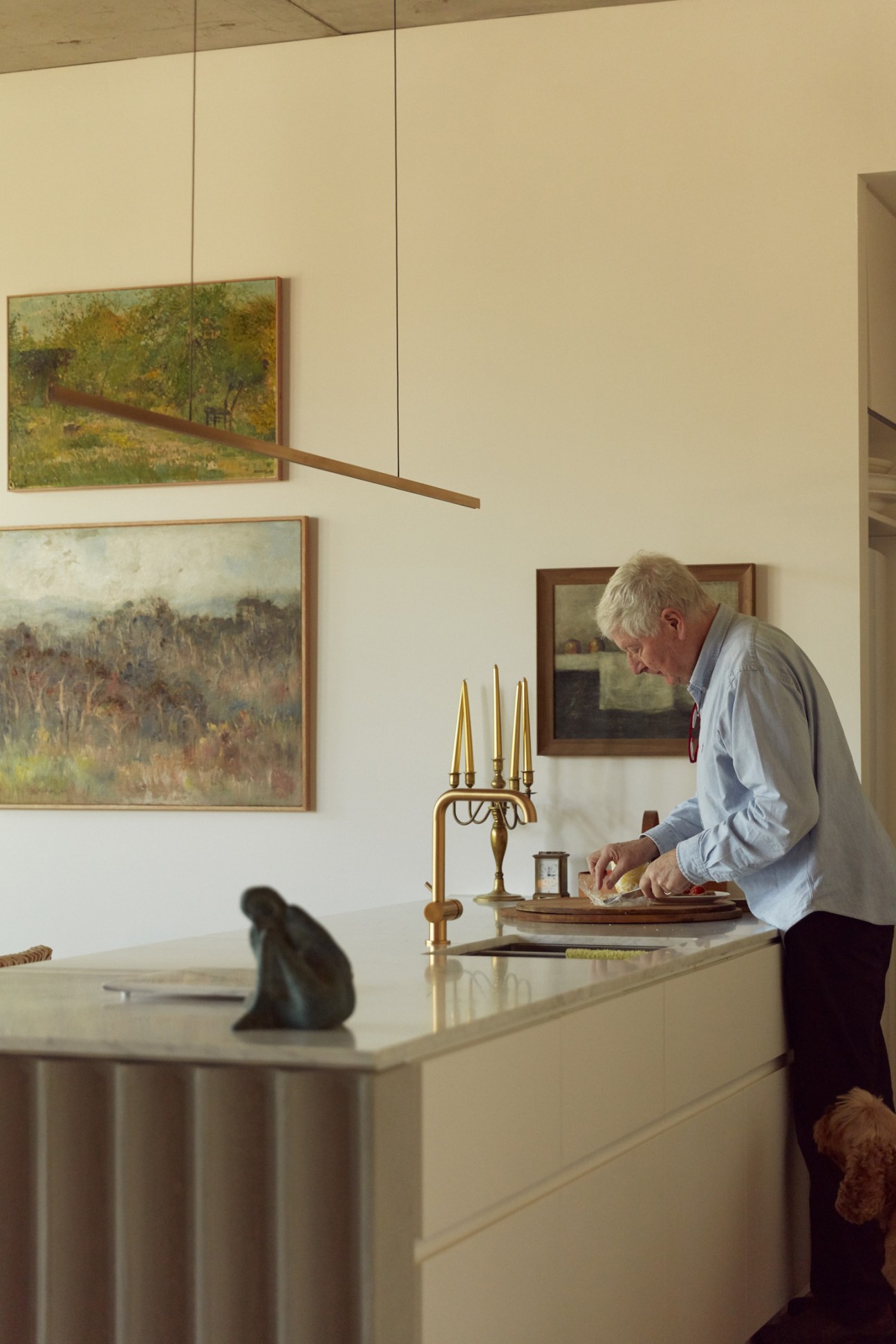Malcolm Good stands at a marble countertop in a minimalist apartment kitchen with framed landscape paintings on the wall.