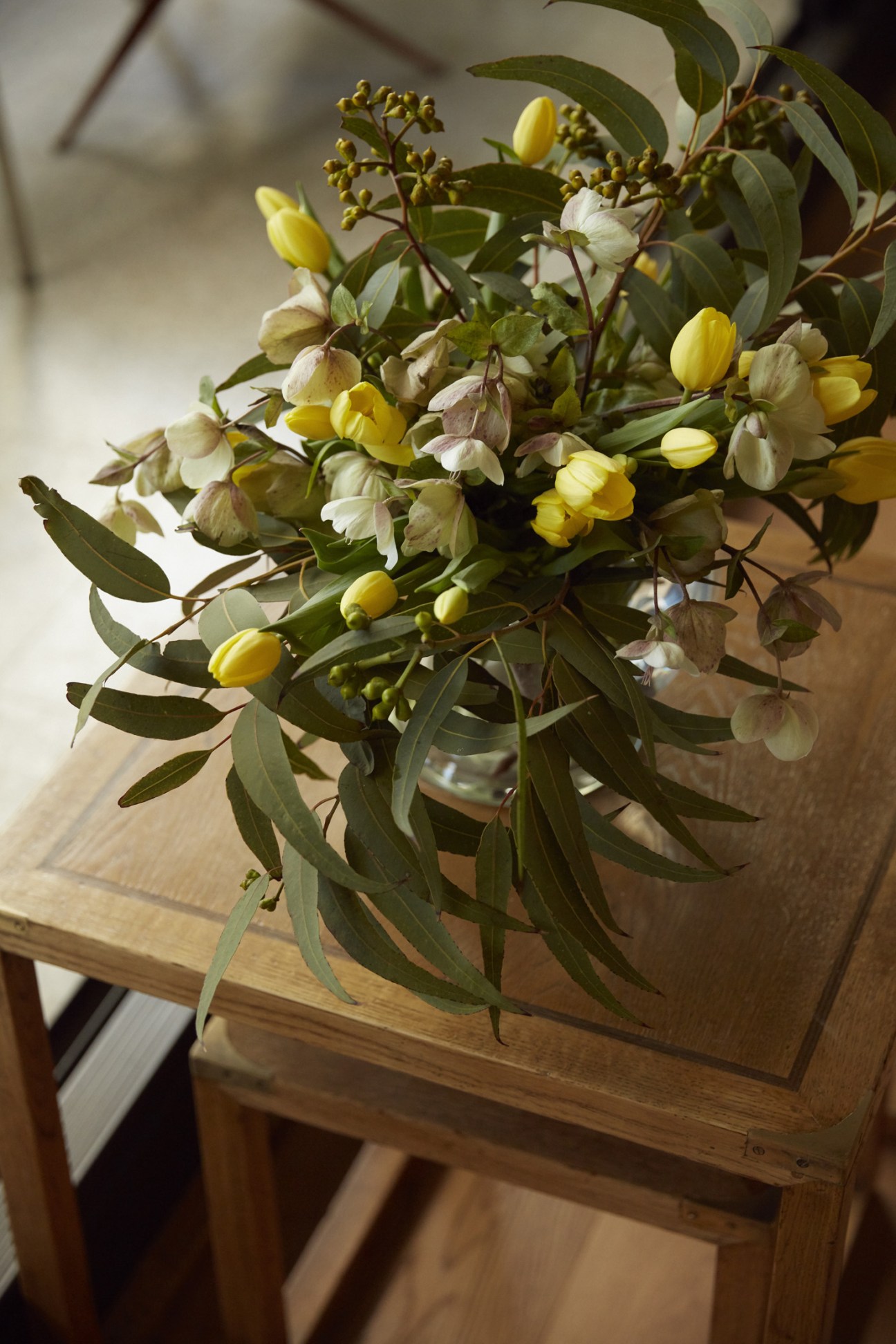 A floral arrangement with yellow and white flowers and green eucalyptus leaves in a vase on a wooden table.