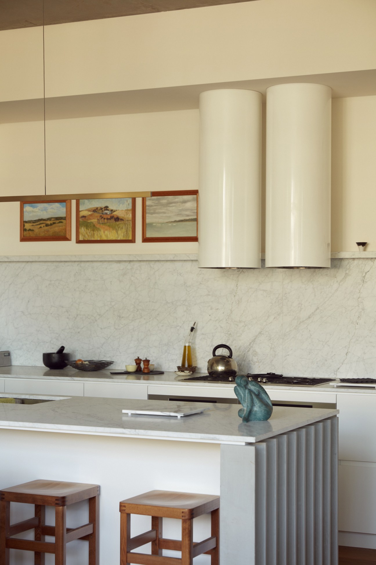 Minimalist kitchen with Carrara marble countertops, white cabinetry, and framed landscape artwork on cream walls.