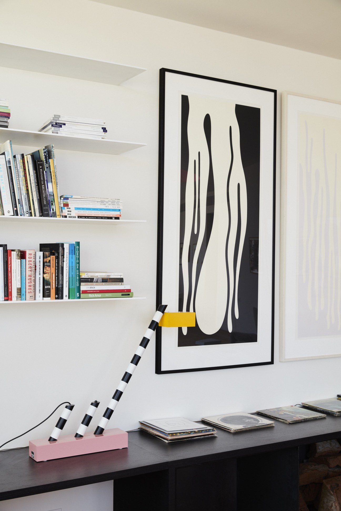 Interior detail showing framed abstract artwork, white shelving with books, and a sculptural object with black and white striped segments on a desk.