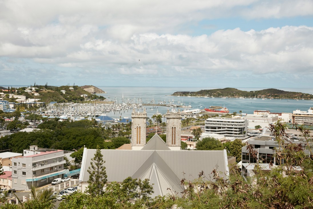 Aerial view of Nouméa's waterfront with marina, church spires, and tropical vegetation overlooking the bay.