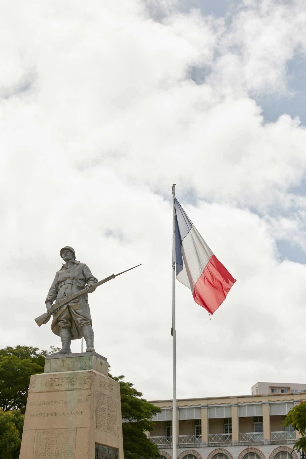 Monument Aux Morts statue of a soldier holding a rifle next to a French tricolour flag under cloudy sky.