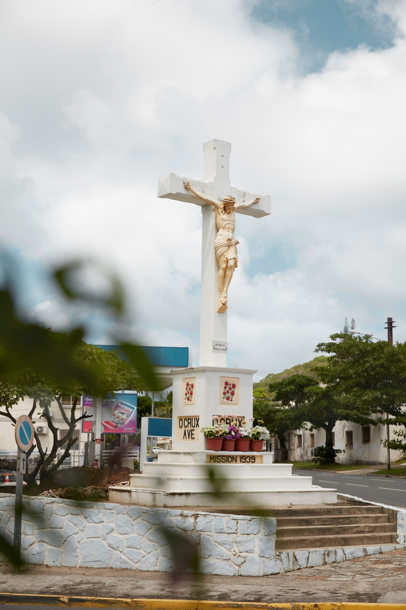 White crucifix monument with golden Jesus figure, base labeled 'Mission 1939', surrounded by tropical trees and buildings.