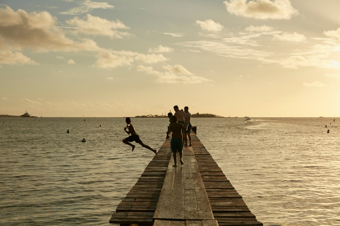 Young people running and jumping off a wooden pier into calm ocean waters at sunset.