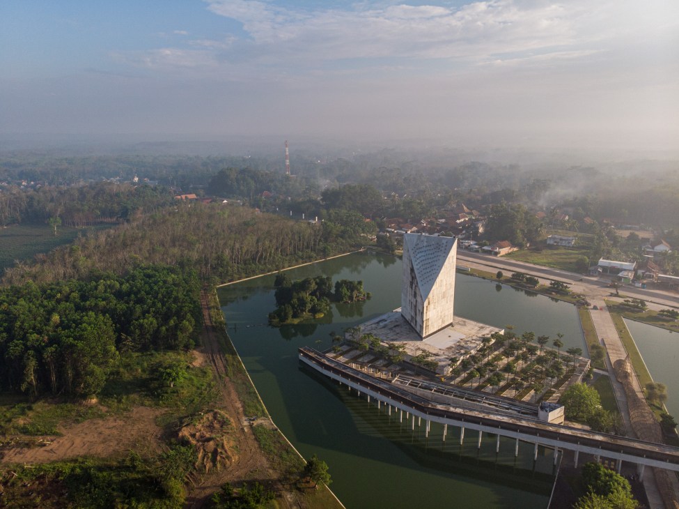 Aerial view of As-Sobur Mosque's white concrete tower rising from jungle landscape in Tulang Bawang Barat, Indonesia, surrounded by water and forest.