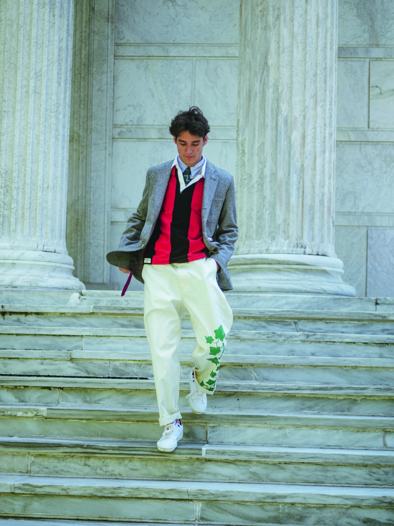 Man wearing light blue blazer, red and black sweater, white pants with green leaf print, and white sneakers on stone steps.
