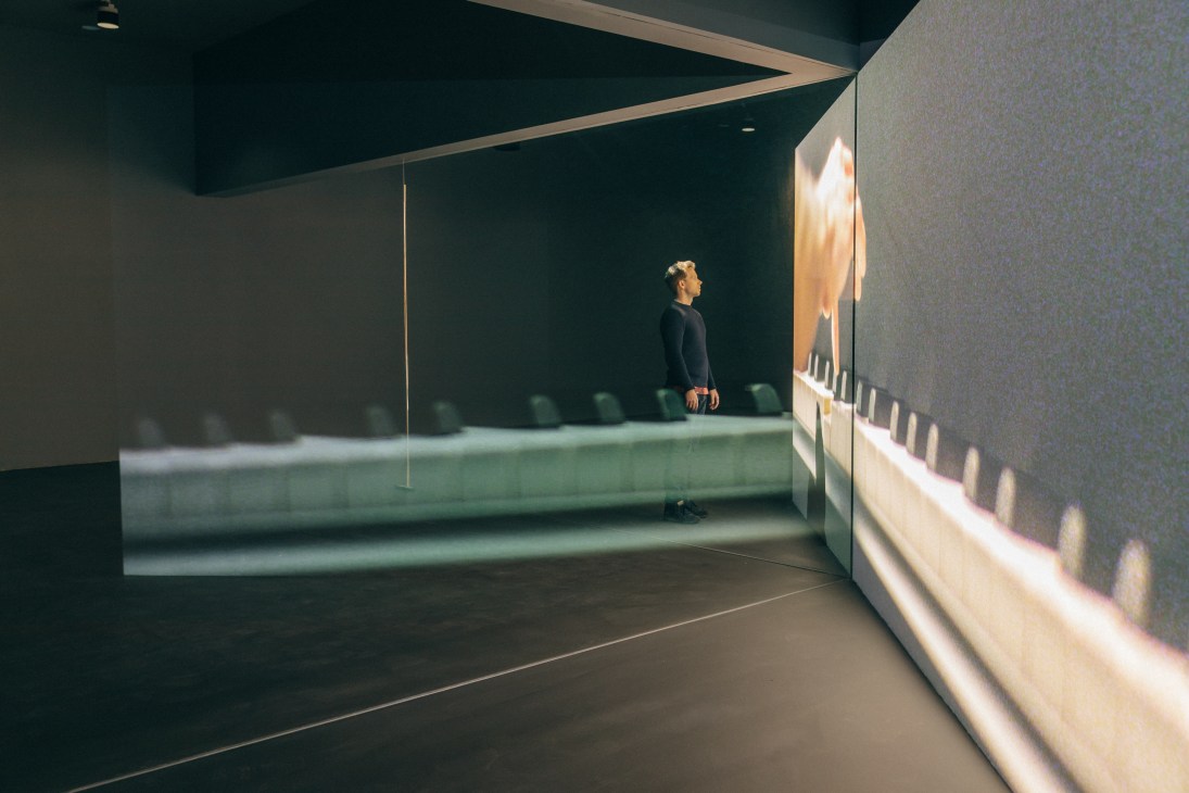 A person stands in a gallery space viewing Anri Sala's video installation 'Take Over' on illuminated screens.