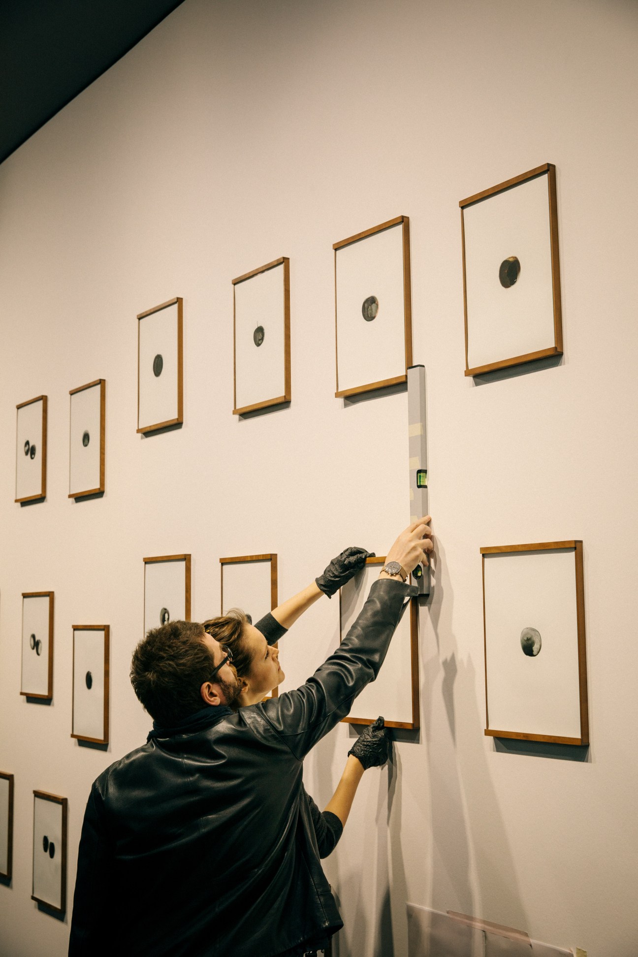 Gallery assistant adjusting a framed artwork on a white gallery wall with multiple minimalist framed pieces.