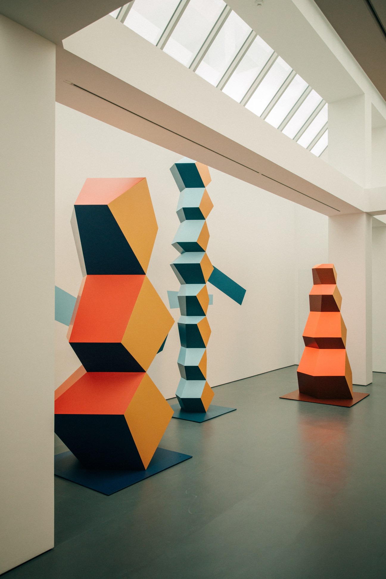 Colorful geometric sculptures displayed in a minimalist gallery space with skylights.