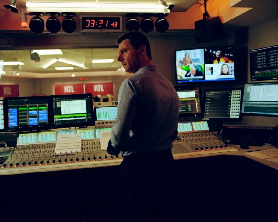 Man in control room at RTL radio station with mixing console, monitors, and red RTL branding visible.