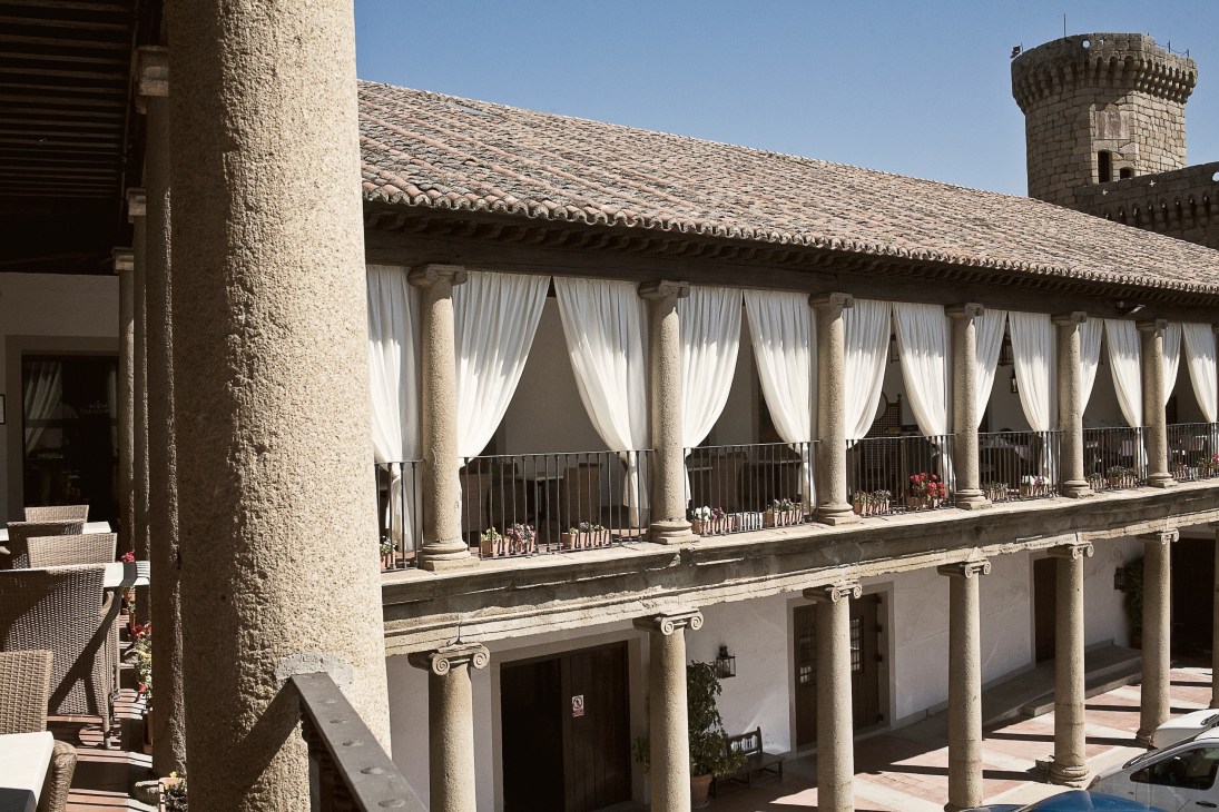 Colonnade with white curtains and stone columns overlooking a courtyard with a tower in the background.