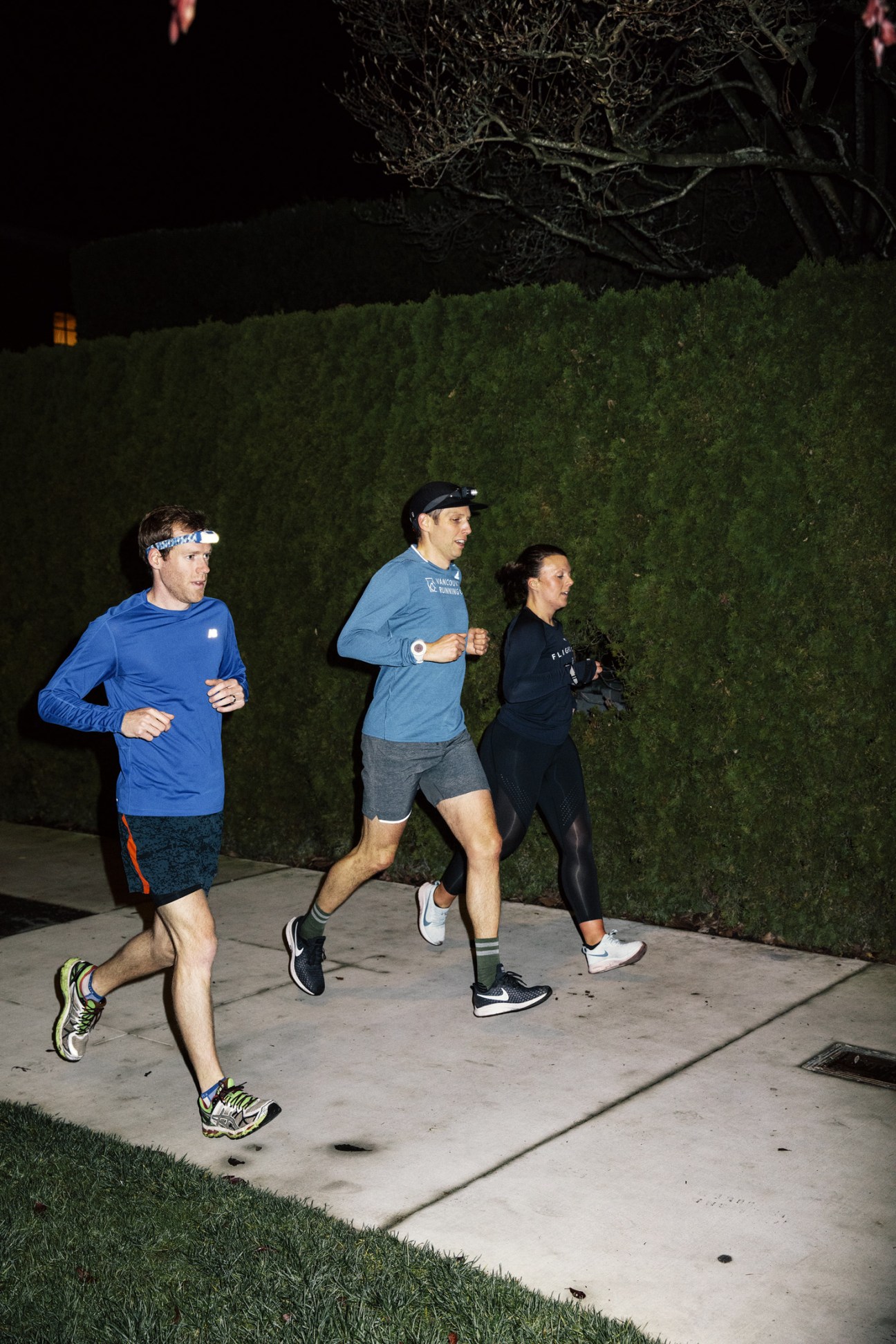 Three runners in technical gear running at night on a sidewalk past hedges.