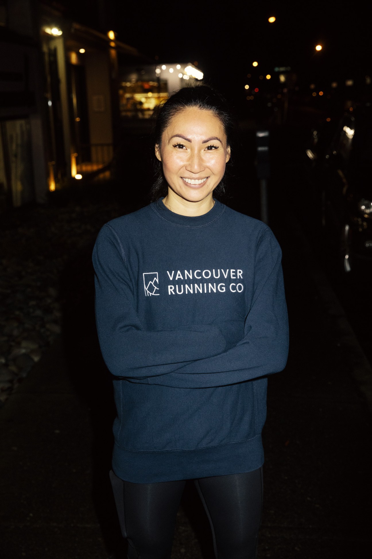 A smiling runner wearing a navy Vancouver Running Co sweatshirt stands on a dark street at night.