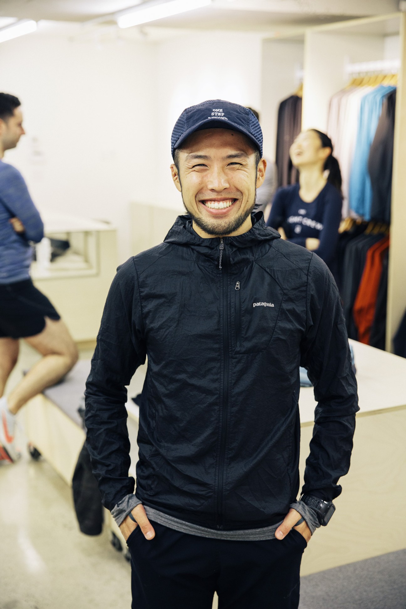 A smiling man wearing a navy Patagonia jacket and cap stands inside Vancouver Running Co store with other runners in the background.