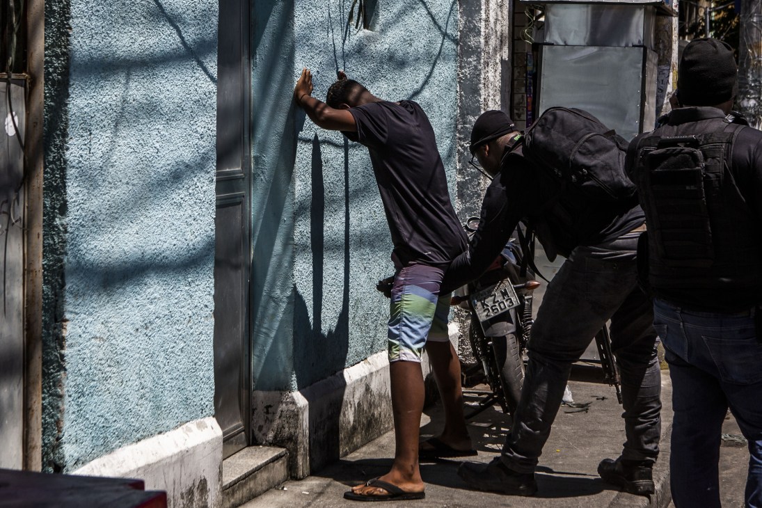 Military police officers frisk a man against a light blue wall during a drug-trafficking operation in Rio de Janeiro's Cruzeiro favela.