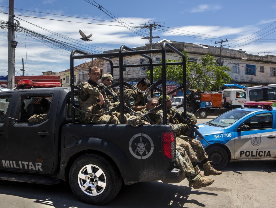 Armed military police troops in camouflage uniforms sit in the back of a black pickup truck marked 'MILITAR' in a favela, preparing for deployment.