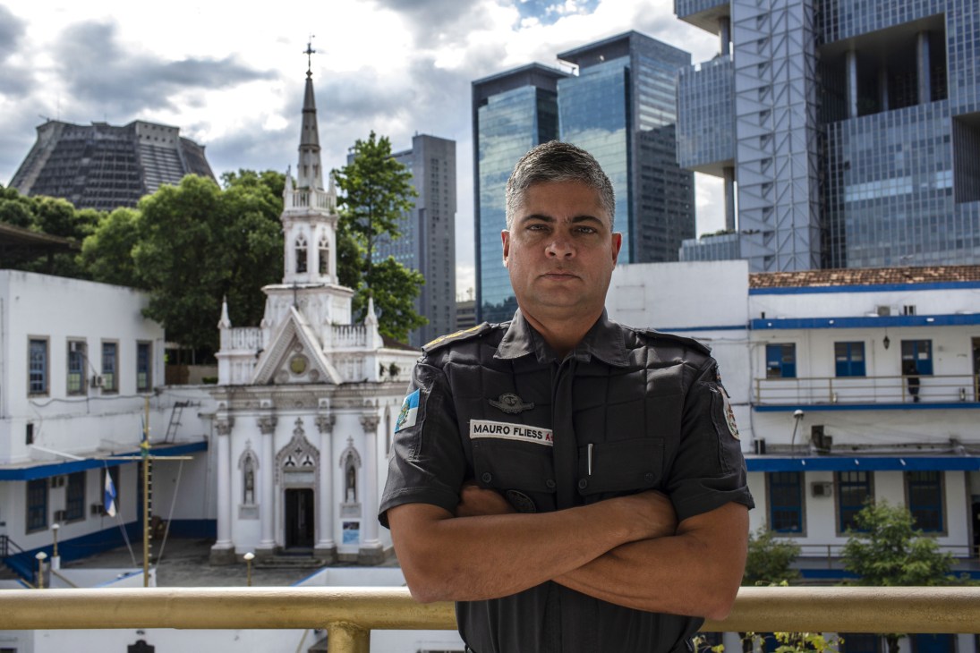 Colonel Mauro Fliess, Rio de Janeiro's Military Police spokesman, stands with arms crossed wearing a black military uniform against a Rio cityscape.