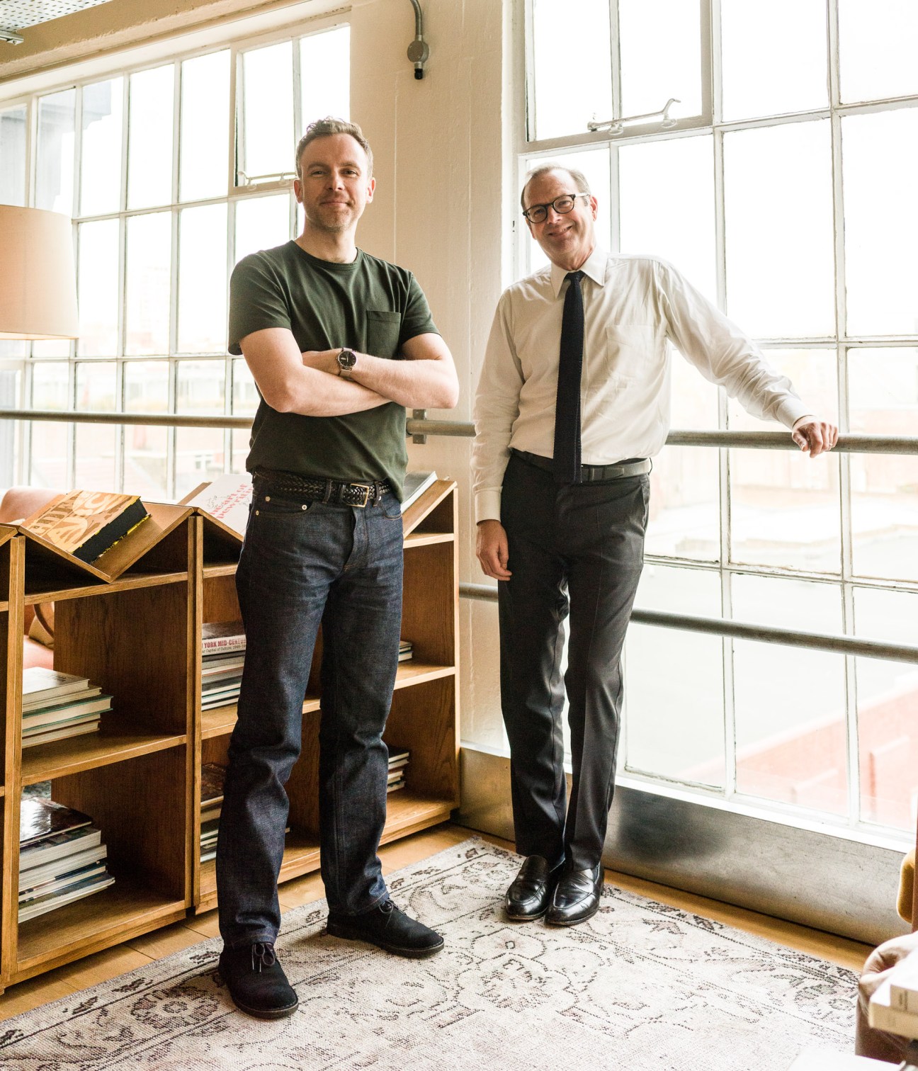 Two men stand in a bright studio space with large windows, next to wooden shelving with books.