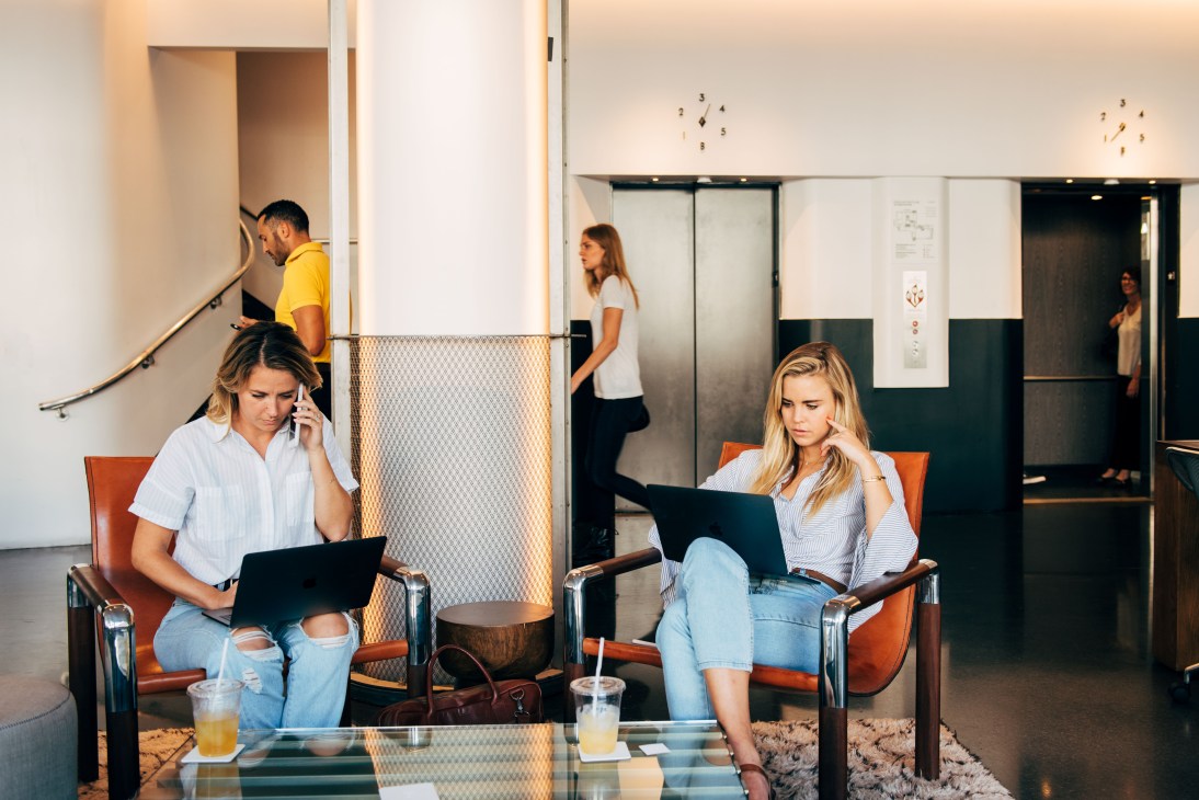 Two women work on laptops in orange leather chairs at NeueHouse co-working space.