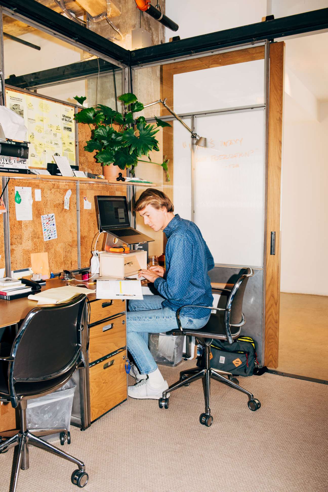 Person working at a laptop in a private desk cubicle with glass partitions and cork board.