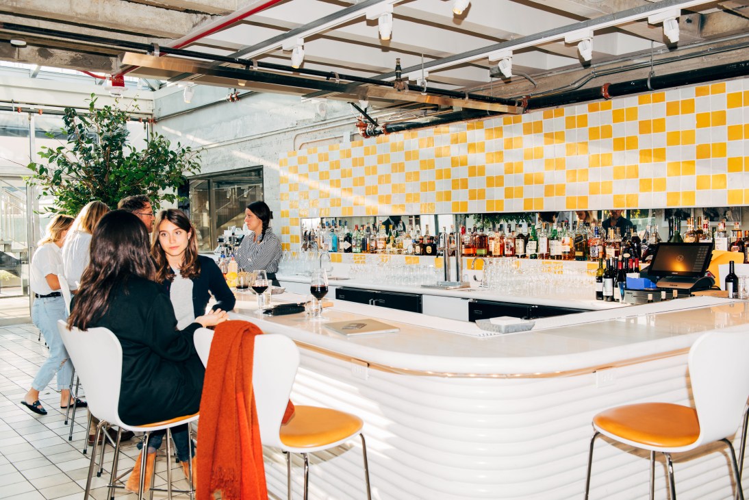 People socializing at a modern bar with yellow and white checkered wall tiles and well-stocked shelves.