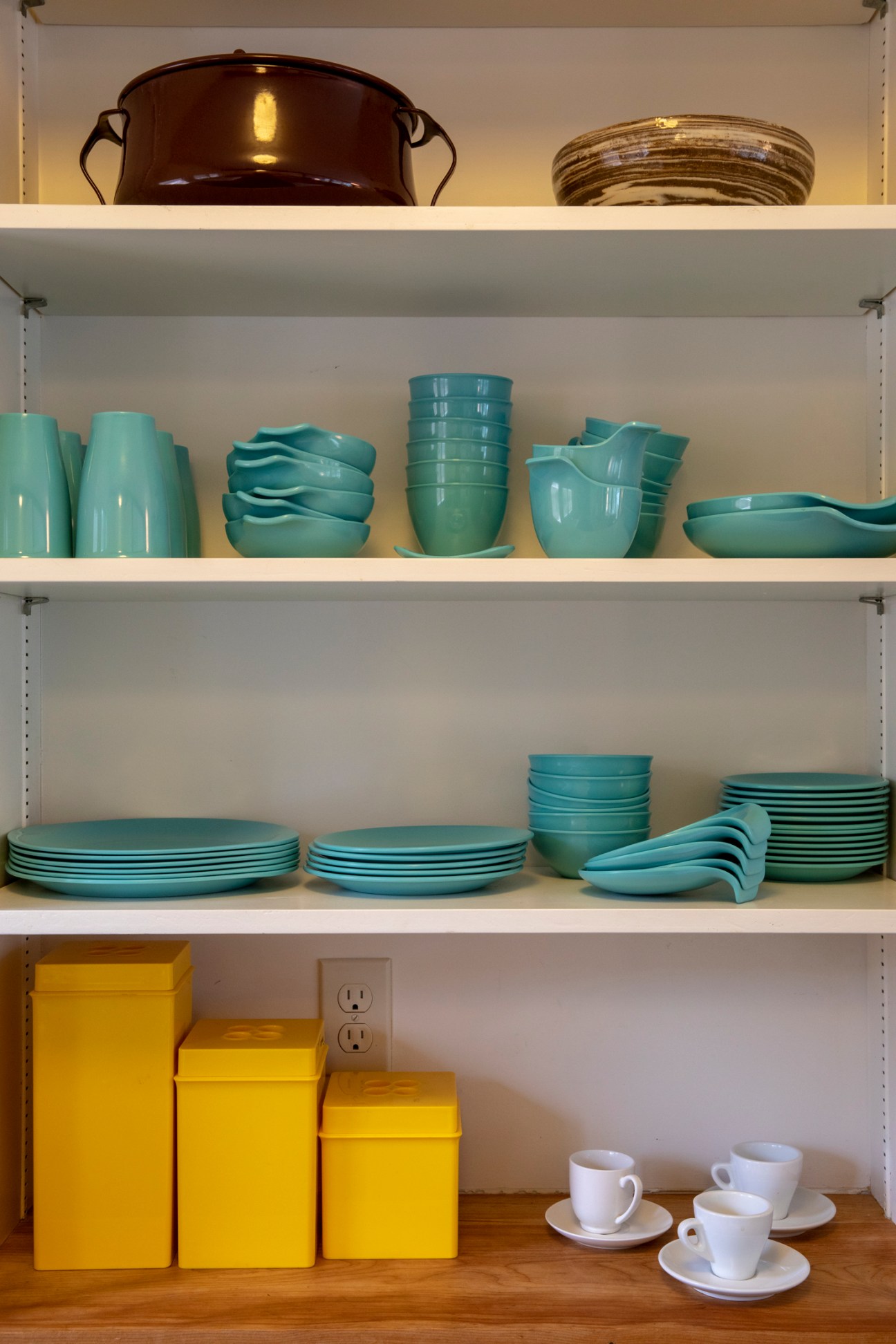 Kitchen shelving with turquoise dishware, yellow storage boxes, white cups and saucers displayed on white shelves.