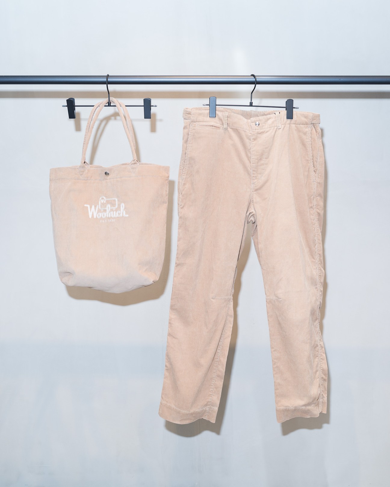 Beige Woolrich tote bag and matching chinos hanging on a rail with black clips.