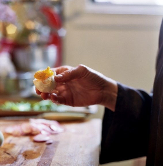 A hand holding a boiled egg with golden yolk visible, with kitchen ingredients blurred in background.