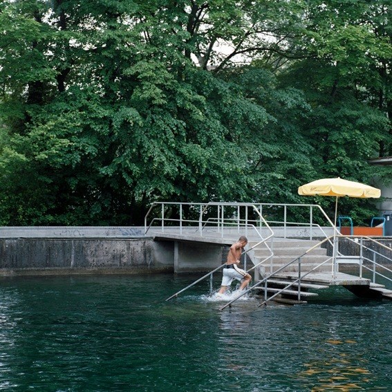 Person swimming in a river from a metal dock with yellow umbrella under lush green trees.