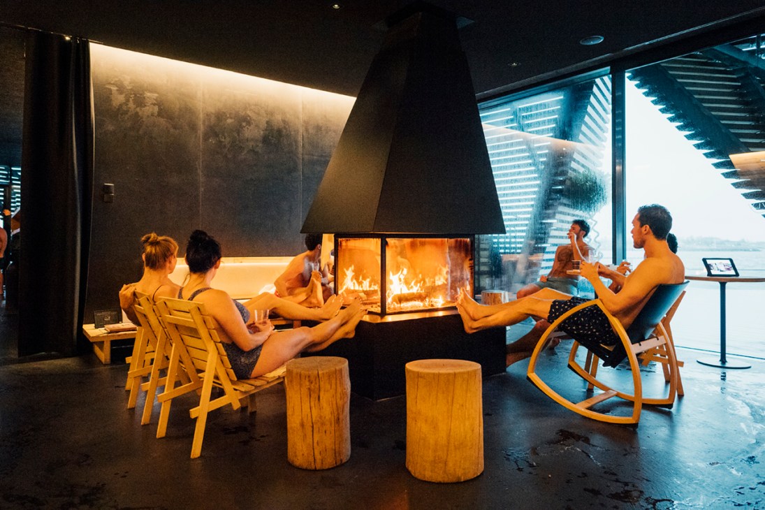 People relaxing in wooden chairs by a fireplace at Löyly sauna in Helsinki with a sea view.