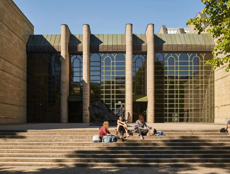 People sitting on steps in front of a modern building with large columns and arched windows in Munich's art district.