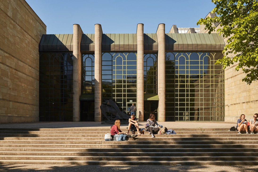 People sitting on steps in front of a modern building with large columns and arched windows in Munich's art district.