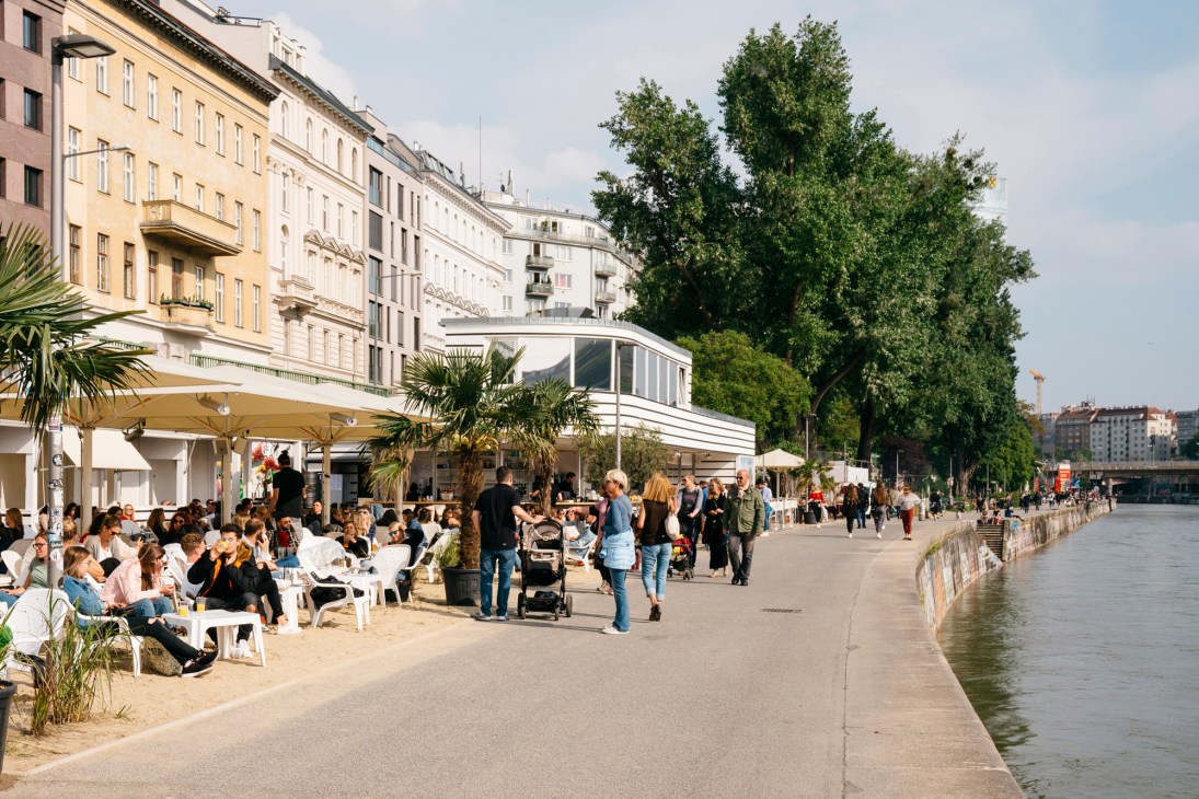 Crowded waterfront bar with sandy seating area along Vienna's Danube Canal, featuring palm trees and classical buildings.