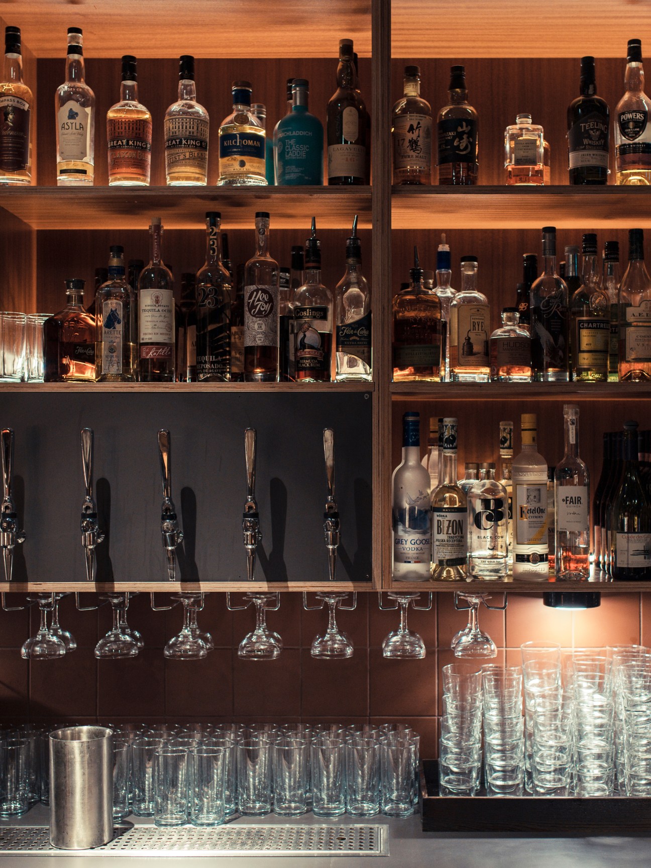Wooden shelving displaying bottles of spirits and whiskey with pull taps and hanging glassware below.