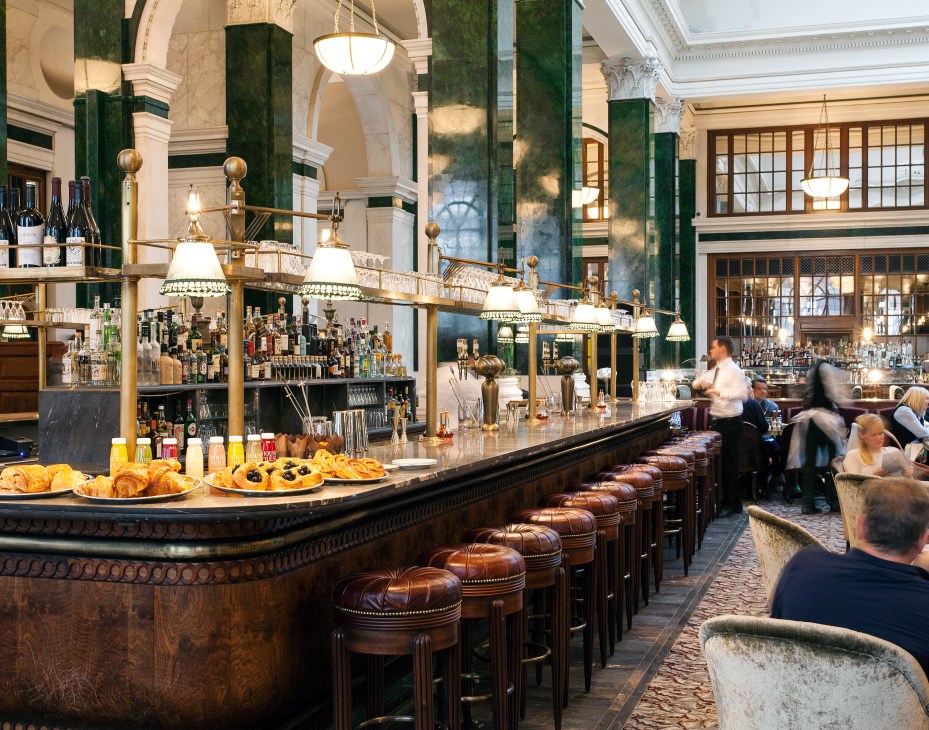 A luxurious restaurant interior with green marble columns, brass fixtures, bar seating, and patrons dining beneath ornate lighting.